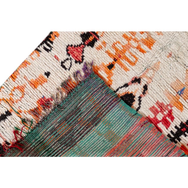 A vintage Moroccan rug with an ivory field and a vibrantly-colored tribal design of orange, red, blue and yellow. This...