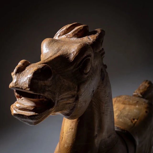 Large carved wooden carousel horse with an iron stand. The carved natural wood surface with visible grains gives it a...