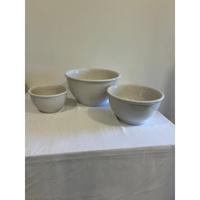 A classic set of three vintage white stoneware mixing bowls in graduated sizes. The bowls feature substantial rolled rims,...