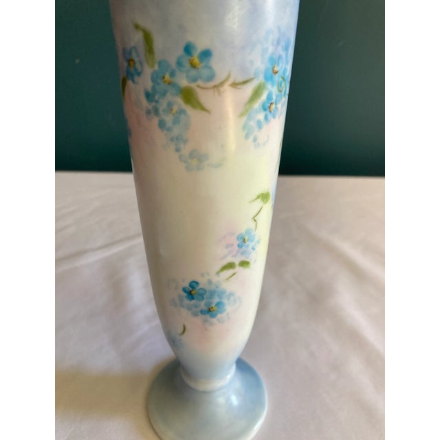 This charming small pedestal vase from the 1920s features delicate hand-painted blue floral motifs on a soft white...