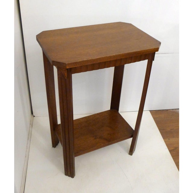 A small, two-tiered side table (pedestal table) in the Art Deco style, with a top shelf featuring chamfered corners and a...