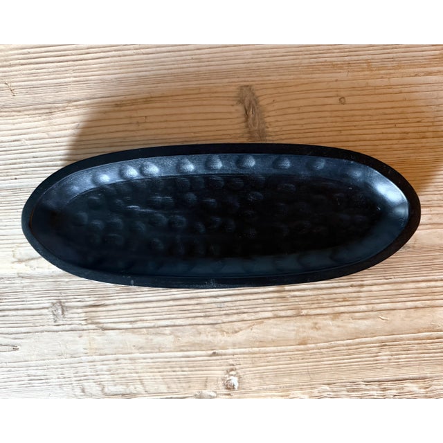 An elongated black wood tray featuring a distinctive hammered-style texture and streamlined oval form. The matte finish...