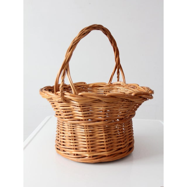 This vintage wicker basket features a rounded woven body, gently flared rim, and tall arched handle in a warm honey-toned...