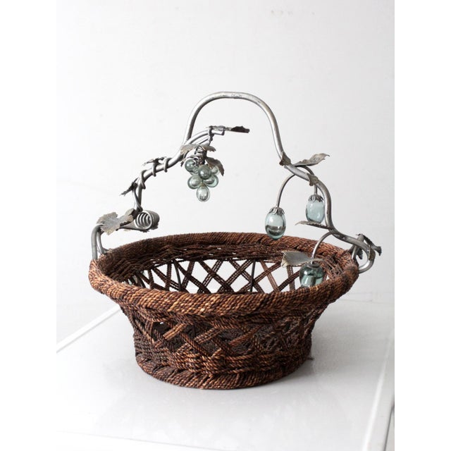 This large vintage basket features a handwoven base paired with a sculptural metal grapevine handle accented with leaf...