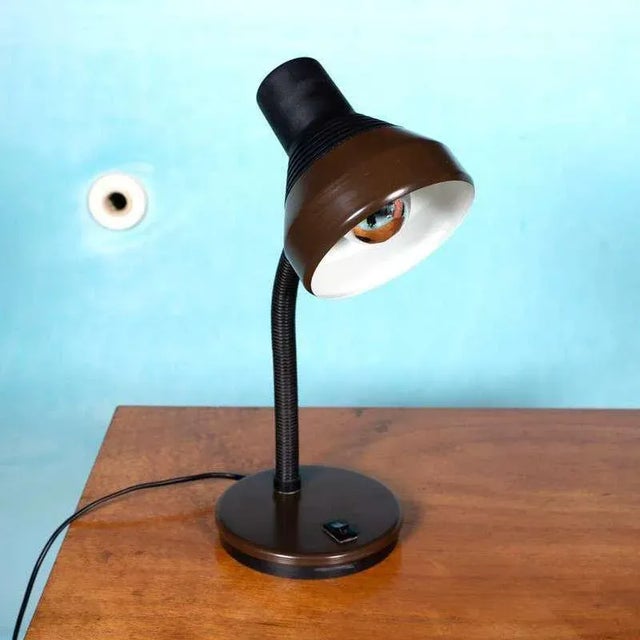 A stylish, sleek desk lamp from the 1970s. The lamp has a round, dark brown metal base with an integrated switch. A...
