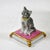 A stunning gray and white cat with yellow collar sitting regally atop a gold border and tasseled pink pillow figurine....