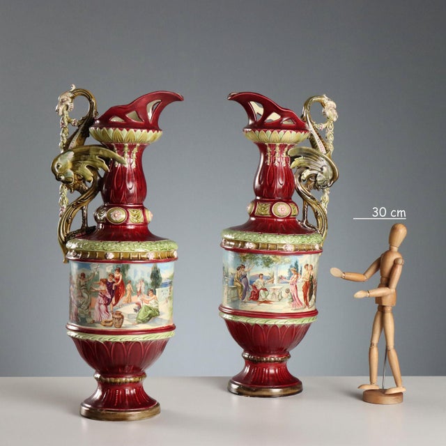 Pair of polychrome ceramic jugs. The body features a band with classical-style decal scenes. The handle is shaped like a...