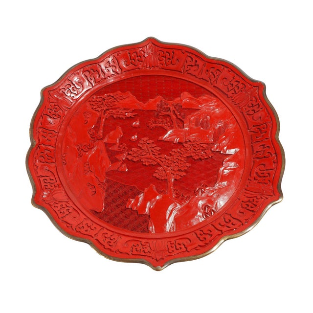 Hand carved cinnabar plate with scalloped rim on brass. The carved scene is of a mountain and river landscape. In the...