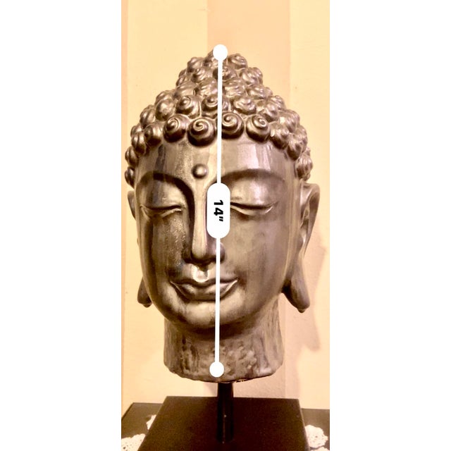 This item is an extra large, ceramic Buddha head sculpture mounted on a metal stand. It is a decorative piece inspired by...