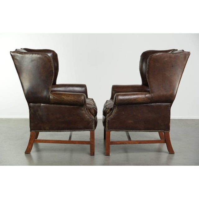 This unique set of two large, characterful English-style cowhide wingback chairs features a beautiful dark cognac color...