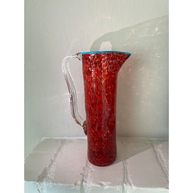 Colorful hand-blown glass pitcher with a red spatter/splatter pattern and turquoise rim, dating to approximately the...