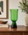 Striking vintage hand-blown art glass pedestal vase in rich emerald green, featuring a tall cylindrical body with a subtly...