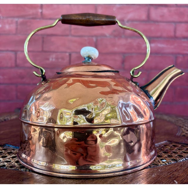 This is a vintage hammered copper tea kettle featuring a rounded body, brass-tone spout and swing handle, wooden grip, and...