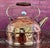 This is a vintage hammered copper tea kettle featuring a rounded body, brass-tone spout and swing handle, wooden grip, and...