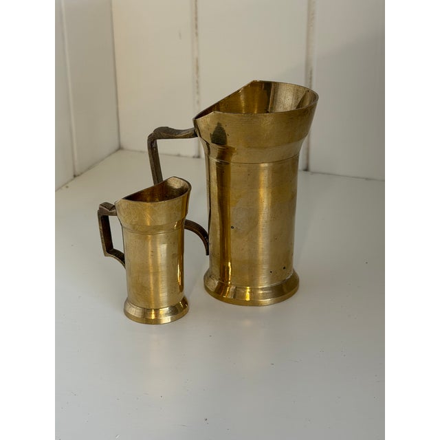 A charming pair of antique brass creamers featuring elegant tapered forms, rolled pouring lips, and angular handles....