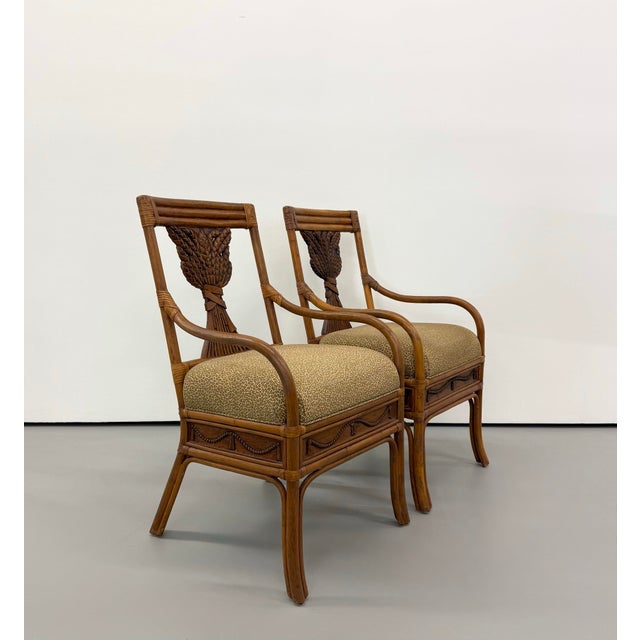 A decorative pair of vintage rattan armchairs featuring sculptural open backs centered by woven wheat sheaf motifs. The...