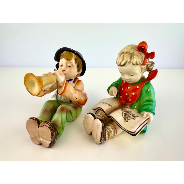 A charming matched pair of mid-century Japanese ceramic figurines depicting a seated boy playing a horn and a young girl...