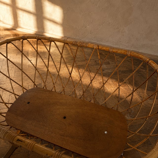A vintage rattan baby bassinet from the 1970s, designed with an oval wicker basket and a low wooden stand. The decorative...