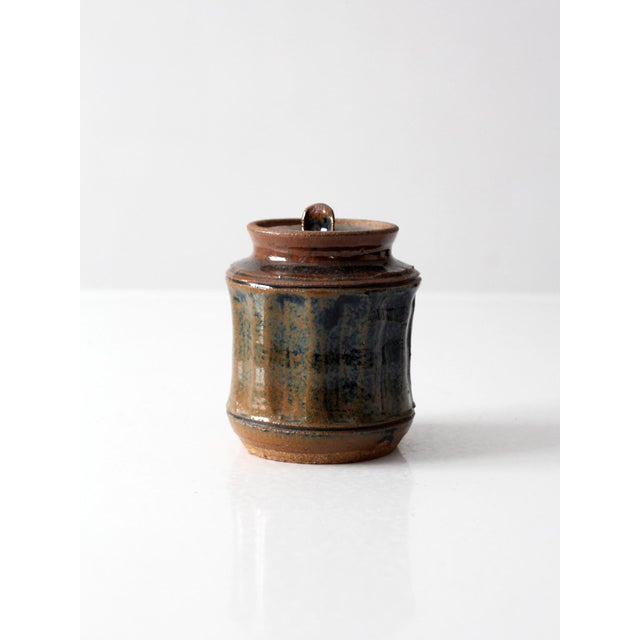 A vintage handmade studio pottery jar with a compact cylindrical form and fitted lid. The surface shows layered blue,...