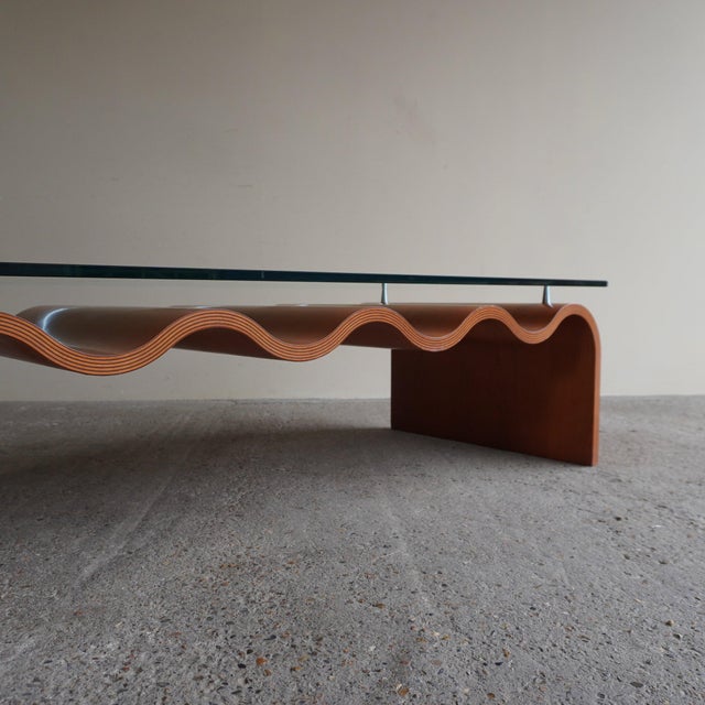 Low table with a steam-bent laminated wood base in a warm terracotta finish, shaped into a continuous undulating wave....