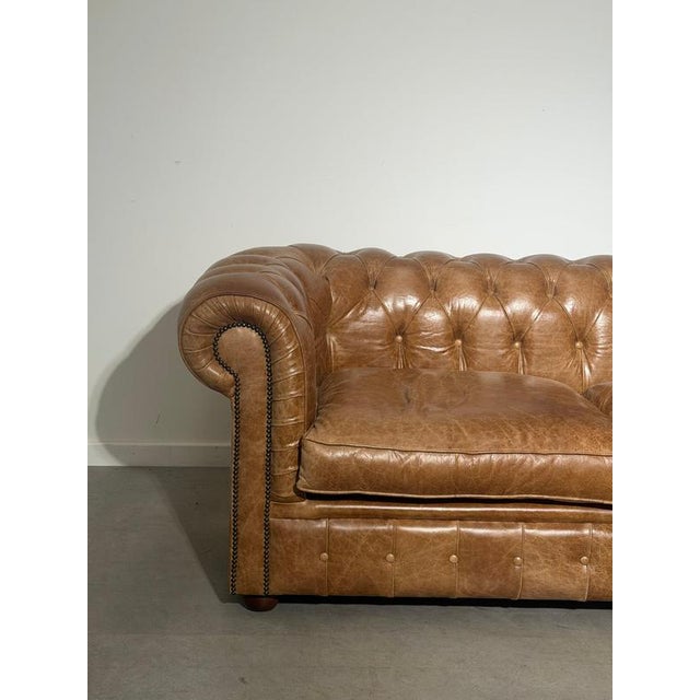 Classic Chesterfield three-seater sofa upholstered in beautifully aged cognac brown leather. A timeless piece featuring...