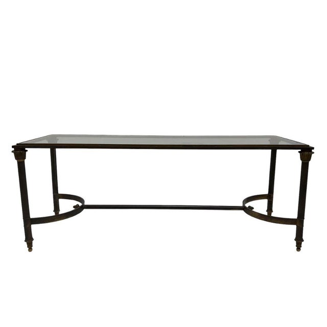 A Neoclassical coffee table crafted with a dark metal frame and clear glass top. The narrow rectangular tabletop is...