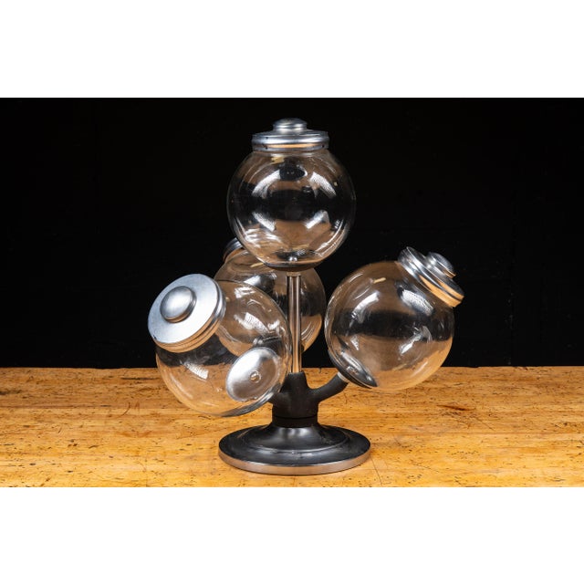 Countertop merchandising display featuring four spherical clear glass storage jars mounted on a revolving metal stand. The...