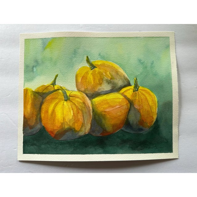 This watercolor painting beautifully depicts a cluster of golden pumpkins bathed in soft light, set against a gentle green...
