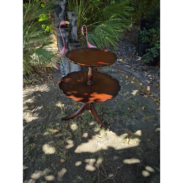 An early 20th-century mahogany two-tier dumbwaiter table features distinctive scalloped "pie-crust" edges and a pedestal...