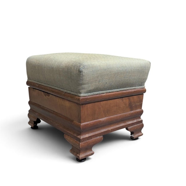 A compact early-to-mid-20th-century storage ottoman with a padded hinged lid, muted green-gray upholstery, and a warm wood...