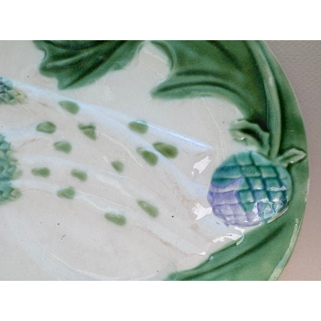 A plate that can be used for asparagus or artichokes. It has raised green leaves and purple tips against a cream...