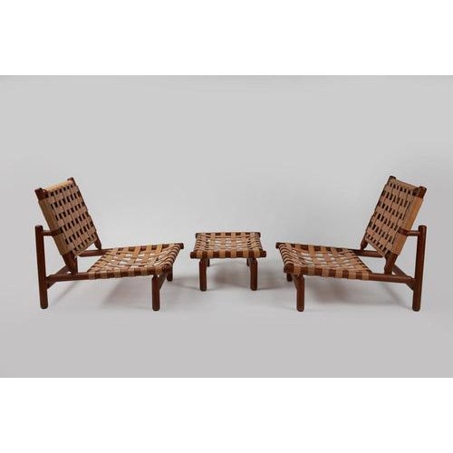 A pair of armchairs with a footstool, made of teak wood, featuring leather straps to support the seat and back cushions....