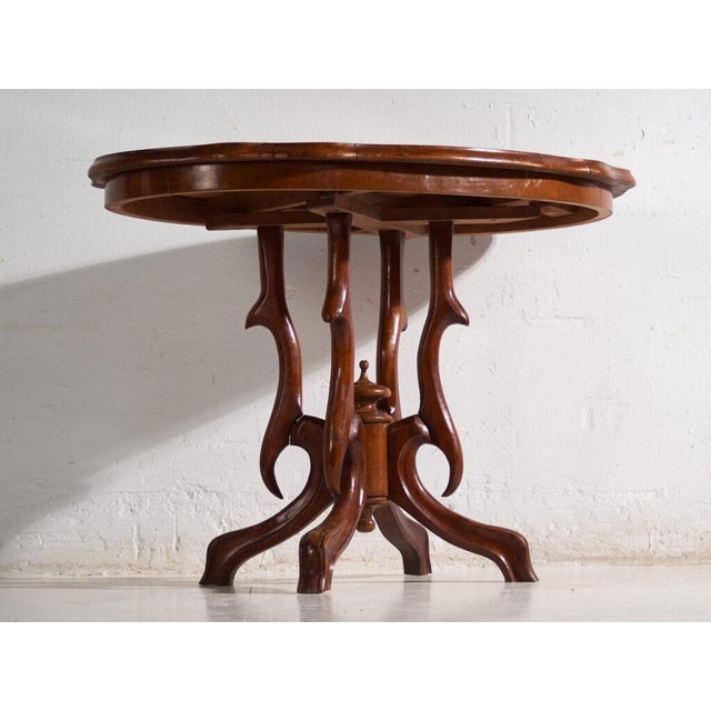 This antique salon table in walnut wood presents an oval top with a shaped outline over a four-armed central pedestal....