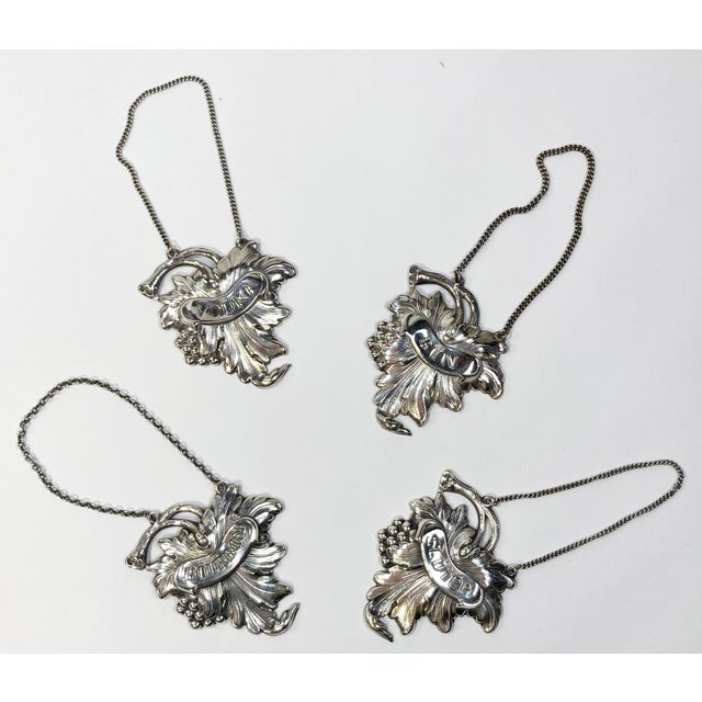 Set of four silver plate liquor tags in a grape and leaf motif. Engraved with GIN, VODKA, SCOTCH and BOURBON. Extends to...