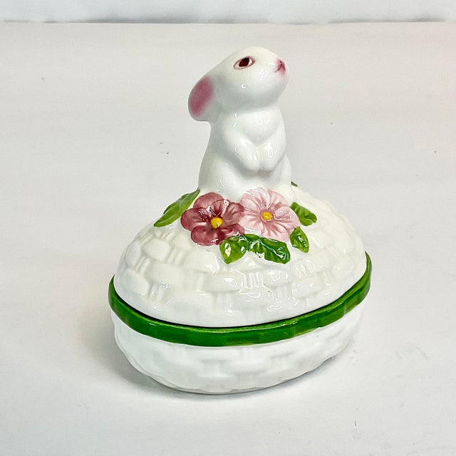 This little bunny will warm your heart. He is sitting up on top of a white ceramic faux wicker basket in the shape of an...