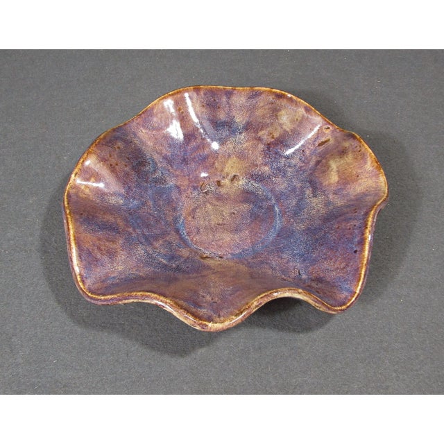 A hand-built studio pottery bowl with a sculptural wavy rim and richly layered glossy glaze. The surface blends lavender,...