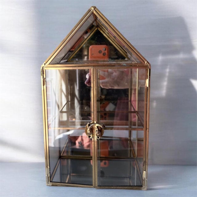 This is a gold-trimmed glass curio cabinet featuring a house-shaped roof and a latching door.This 2-tier display case is...