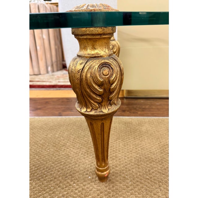 A glamorous cocktail table featuring a thick, curvaceous glass top and four ornate giltwood legs with carved foliate...