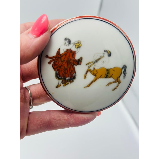 This is a small vintage porcelain trinket (or pill) box decorated in a Neoclassical classical motif. The circular lid is...