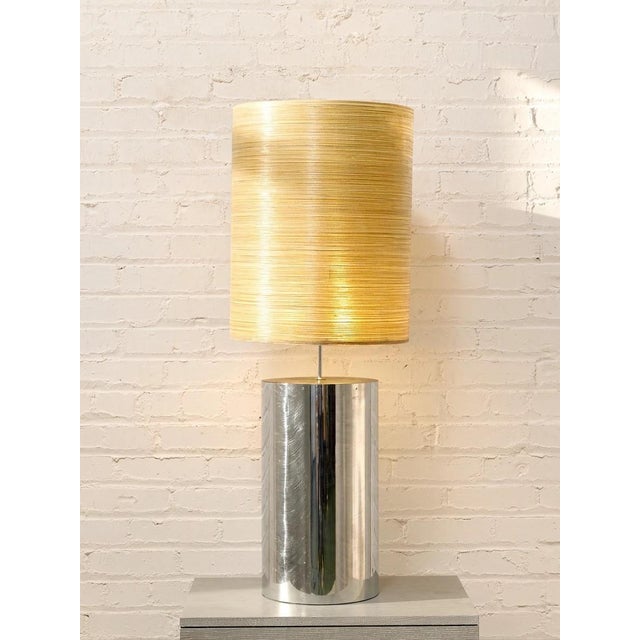 A sleek modern table lamp featuring a polished chrome cylindrical base paired with a tall gold-toned grasscloth shade. The...