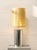 A sleek modern table lamp featuring a polished chrome cylindrical base paired with a tall gold-toned grasscloth shade. The...