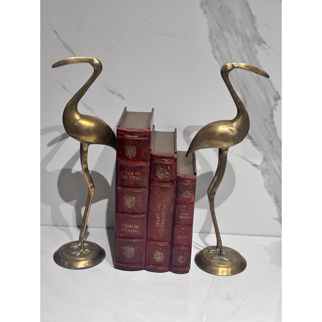 The image features a pair of vintage solid brass crane figurines standing gracefully on round bases with a warm, aged patina.