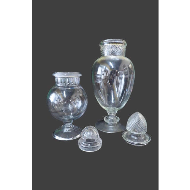 Two large apothecary jars with corresponding lids. Both have a similar bulbous shape to them with a strong round stand....