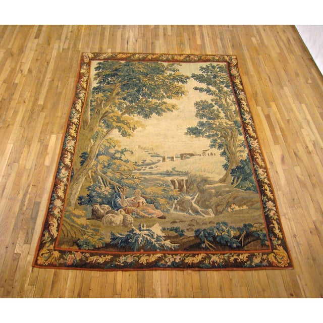 A French pastoral landscape tapestry from the 18th century, depicting a placid country scene with a young couple...