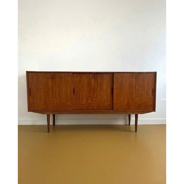 A 1960s danish teak sideboard with three sliding doors and handles carved into the front. The profile is simple and...
