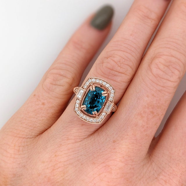 A blue zircon glows in a brilliant electric blue at the heart of this solid 14K rose gold ring, flanked by thirty-two...