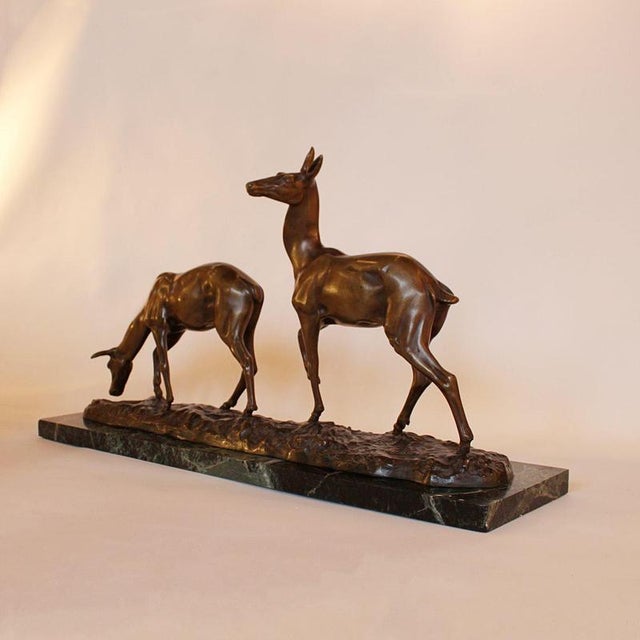 A patinated bronze group of a pair of grazing deer set over a naturalist base on a marble plinth. Signed to bronze....