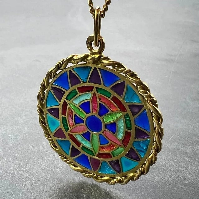 An 18 karat (18K) yellow gold charm pendant designed as a round medal with a rope-twist edge depicting a stained glass...