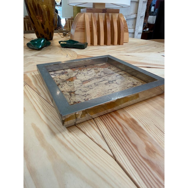 A decorative square tray featuring an organic cork composition beneath a clear glass surface, framed by a broad, silver-...