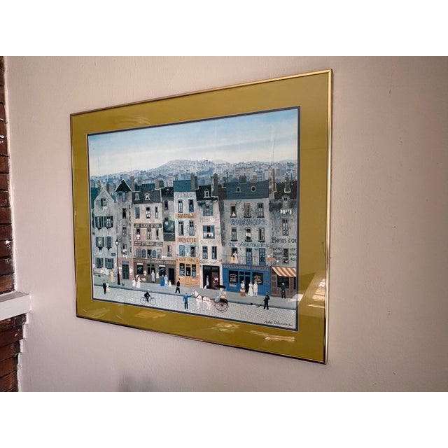 Large framed limited-edition print by French artist Michel Delacroix depicting a charming Parisian neighborhood filled...